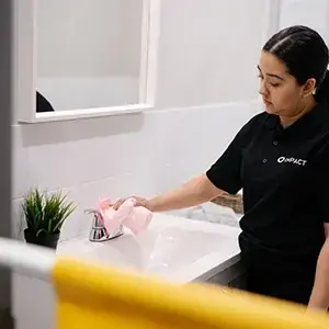 A person in a black polo shirt from Impact Commercial Cleaning Services cleans a bathroom sink with a pink cloth. A small green plant sits on the counter, and a yellow towel hangs in the foreground.