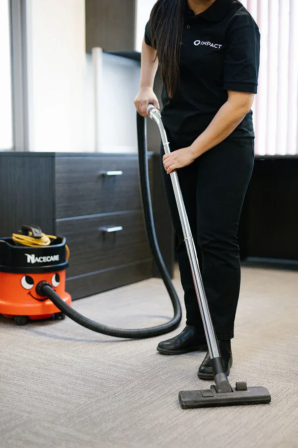 A person in black clothing is vacuuming an office carpet with a red and black commercial vacuum cleaner from Impact Commercial Cleaning Services. Only the person’s torso and lower body are visible, reflecting the professionalism of commercial cleaning Canada.
