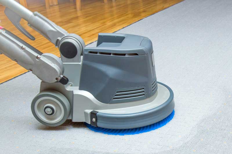 A carpet cleaning machine with a blue brush is scrubbing a light-colored carpet, making the cleaned area visibly brighter than the rest. Wooden flooring is visible in the background, showcasing the impact commercial cleaning services provide.