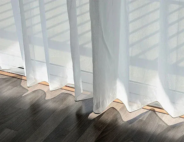 Sheer white curtains hang in front of a window, casting soft shadows and wavy patterns of sunlight onto a wood floor—showcasing the pristine results possible with impact commercial cleaning services.