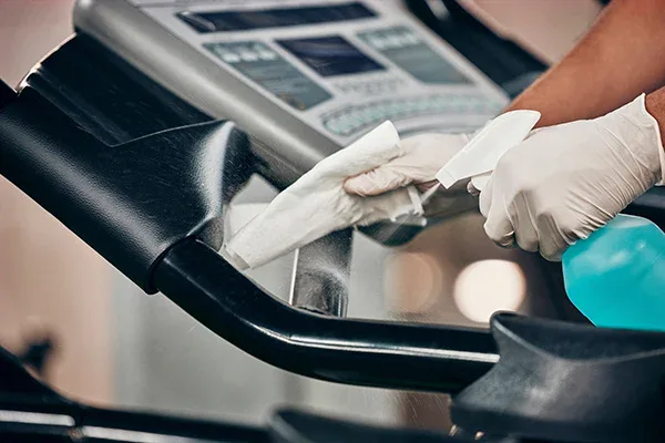 A person wearing white gloves cleans a treadmills handle with a white cloth and sprays blue cleaning solution, ensuring the equipment is sanitized.