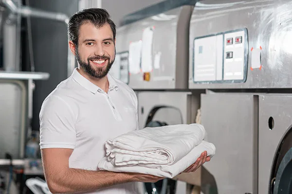 A smiling man in a white polo shirt holds folded laundry in front of industrial washing machines, showcasing the results of impact commercial cleaning services in a busy laundromat or laundry facility.
