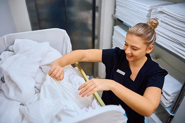 A woman in a black uniform smiles while handling clean white linens from a laundry cart in a room filled with neatly stacked towels and sheets, showcasing the professionalism of impact commercial cleaning services in Canada.