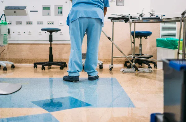 A person in blue scrubs and dark shoes mops the floor of a hospital room, with medical equipment, stools, and supplies visible in the background.