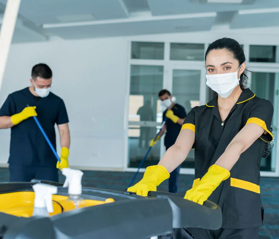 Three people in uniforms and masks are cleaning a large indoor space. Representing impact commercial cleaning services, one woman in front pushes a cleaning cart with spray bottles, while two others mop the floor. All wear yellow gloves.