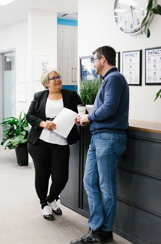 Two colleagues chat in an office near a counter with plants and posters. One holds documents, the other a coffee cup. Both are smiling and dressed in business casual attire, reflecting the professionalism of impact commercial cleaning services.