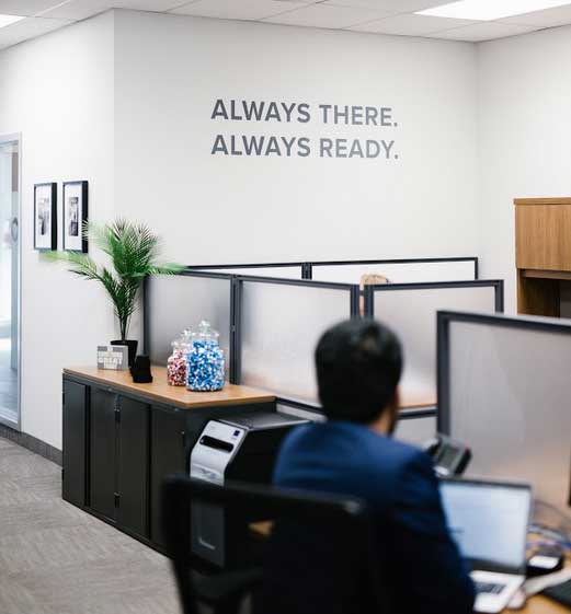 A modern office with cubicles, a person working at a desk, jars of colorful candy on a cabinet, a potted plant, and a wall sign that reads ALWAYS THERE. ALWAYS READY—an ideal space for impact commercial cleaning services.