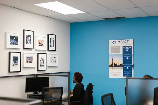 A person sits at a computer desk in a modern office with blue and white walls, framed photos, and an IMPACT poster highlighting impact commercial cleaning services. Several cubicles and office chairs are visible.