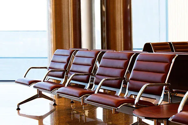Row of empty brown leather chairs with metal frames in an airport waiting area, with large windows and sunlight streaming in.
