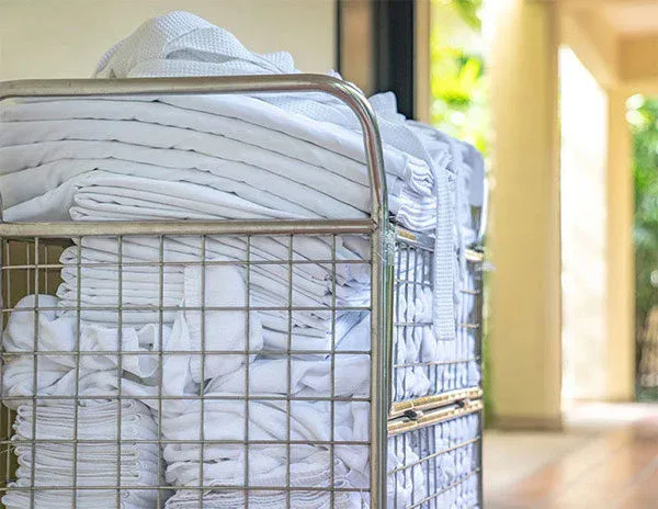A metal laundry cart filled with neatly folded white towels is positioned in a well-lit hallway, highlighting the standards of impact commercial cleaning services, with blurred greenery visible in the background.