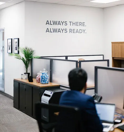 An office with cubicles, a person working at a desk, and jars of colorful candy on a cabinet. The wall displays the words ALWAYS THERE. ALWAYS READY—reflecting the standards of impact commercial cleaning services.