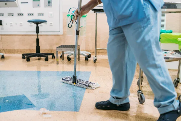 A person wearing blue scrubs and black clogs is mopping the floor in a medical facility or hospital room, with various medical equipment and chairs visible in the background.