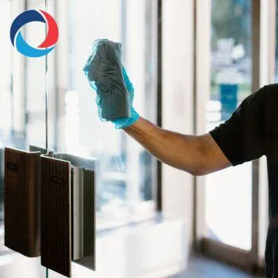 A person wearing a blue glove cleans a glass door with a cloth. Sunlight shines through the windows, and sample panels are attached to the glass. A circular blue and red logo is visible in the top left corner.