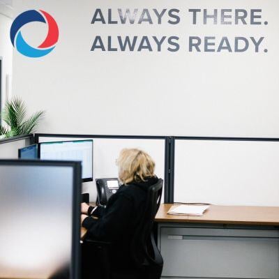 A person with blonde hair sits at a desk in a modern office cubicle, working at a computer. On the wall above is a logo and the text ALWAYS THERE. ALWAYS READY.