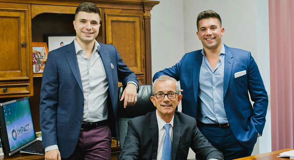 Three men in business attire pose in an office. Two younger men stand on either side of an older man seated at a desk. A wooden cabinet and a computer displaying the word IMPACT are in the background.