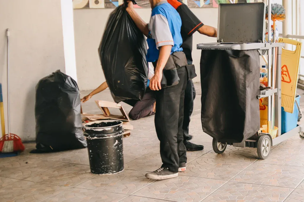 Two people in uniforms are cleaning; one holds a large black trash bag while another reaches for debris on the floor. A cleaning cart with supplies and a bucket are nearby on tiled flooring.