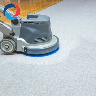A floor cleaning machine with a blue rotary brush is scrubbing a light gray carpet, leaving a visibly cleaner area behind. The machine is on wheels and part of a wooden floor is visible in the background.