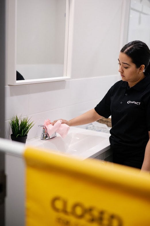 A person in a black shirt cleans a bathroom sink with a pink cloth. A small plant sits on the counter, and a yellow Closed sign highlights the impact commercial cleaning services provided in Canada.