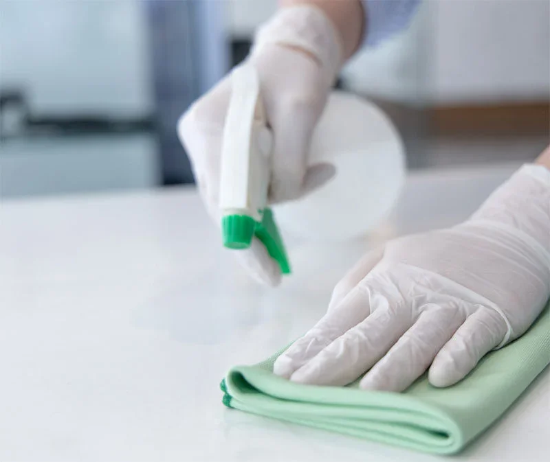 A person wearing white gloves sprays cleaner onto a green cloth while wiping a white surface, illustrating the thoroughness of impact commercial cleaning services.