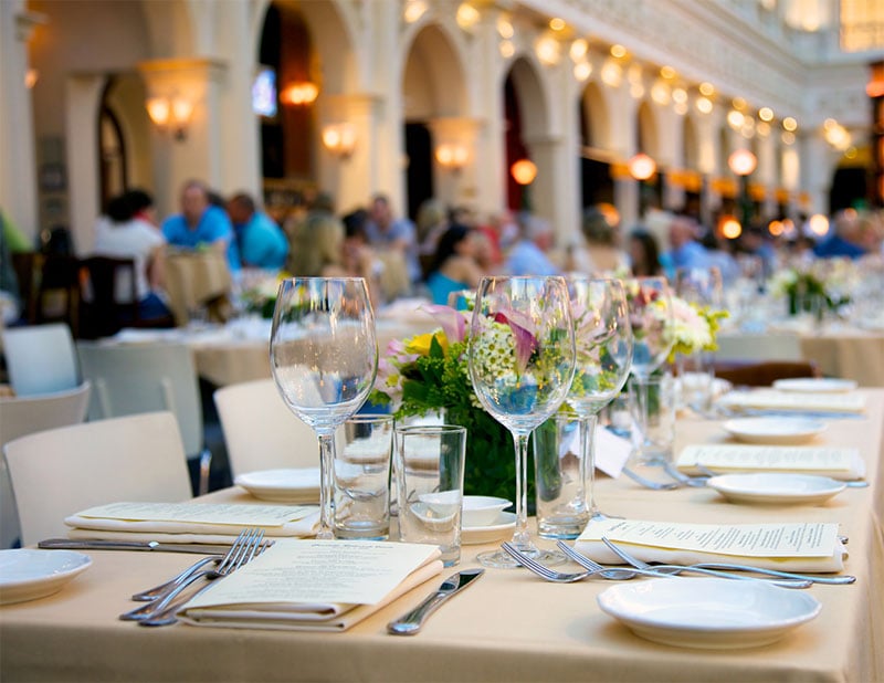 A close-up of an elegant outdoor dining table set with glassware, plates, menus, and floral centerpieces. In the background, people are seated, and a building with arches is softly lit—a setting maintained by impact commercial cleaning services.