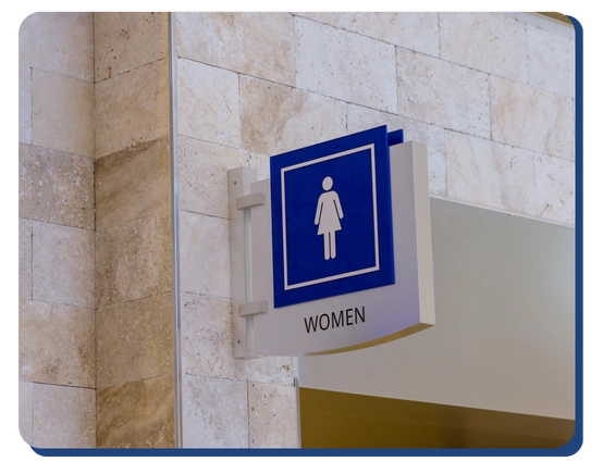 Blue and white restroom sign for women, mounted on a tiled wall above a doorway. Ideal for facilities using Impact Commercial Cleaning Services or commercial cleaning Canada, the sign features a female figure icon and the word WOMEN below it.