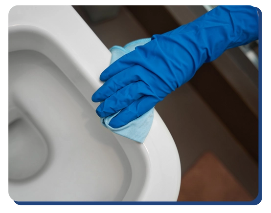 A person wearing blue rubber gloves is cleaning the rim of a white toilet with a light blue cloth.