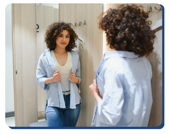 A woman with curly hair tries on a blue and white striped shirt in a fitting room, looking at herself in the mirror with a thoughtful expression, while nearby signage highlights commercial cleaning Canada.