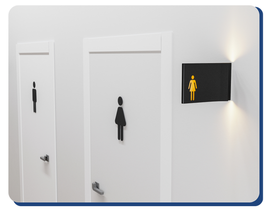Two white restroom doors are shown—one with a male symbol and the other with a female symbol. A black wall sign with a yellow female icon is illuminated above the women’s door, reflecting spaces maintained by Impact Commercial Cleaning Services.