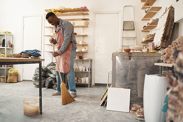 A person wearing an apron sweeps the floor in a ceramics studio filled with pottery materials, shelves with ceramics, and art supplies.