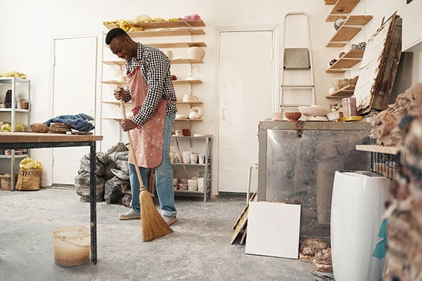 A person wearing an apron sweeps the floor in a pottery studio filled with shelves of ceramics, tools, and clay materials, reflecting the attention to detail found in impact commercial cleaning services.