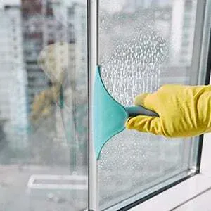 A person wearing a yellow rubber glove uses a blue squeegee to clean a soapy window, with tall buildings visible outside.