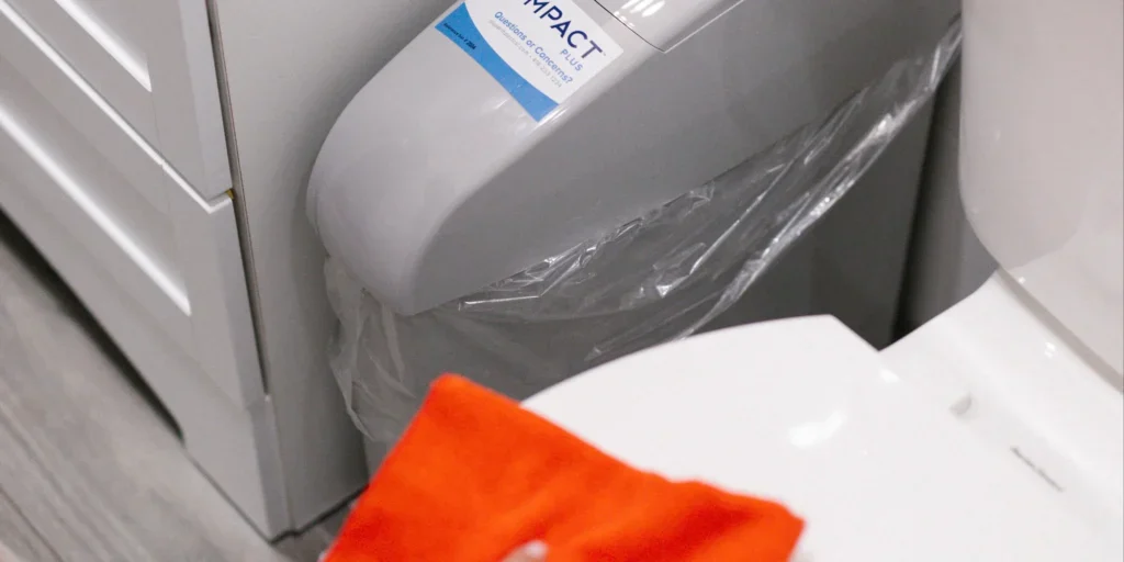 A close-up of a bathroom features a white toilet, a trash bin lined with a plastic bag, and an orange towel on top. A cleaning product labeled IMPACT highlights the professional touch of commercial cleaning Canada services.