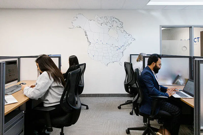Four people working at desks in a modern office, separated by partition walls. A map of North America is displayed on the wall. Everyone appears focused on their computers, reflecting a professional space maintained by impact commercial cleaning services.
