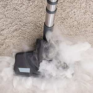 A steam cleaner from Impact Commercial Cleaning Services releases thick steam onto a beige carpet, with the cleaner’s black nozzle and silver handle visible.