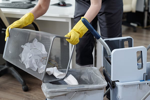 A person wearing yellow gloves empties a mesh trash bin filled with crumpled paper into a larger trash can in an office setting. Cleaning supplies and a desk are visible in the background.