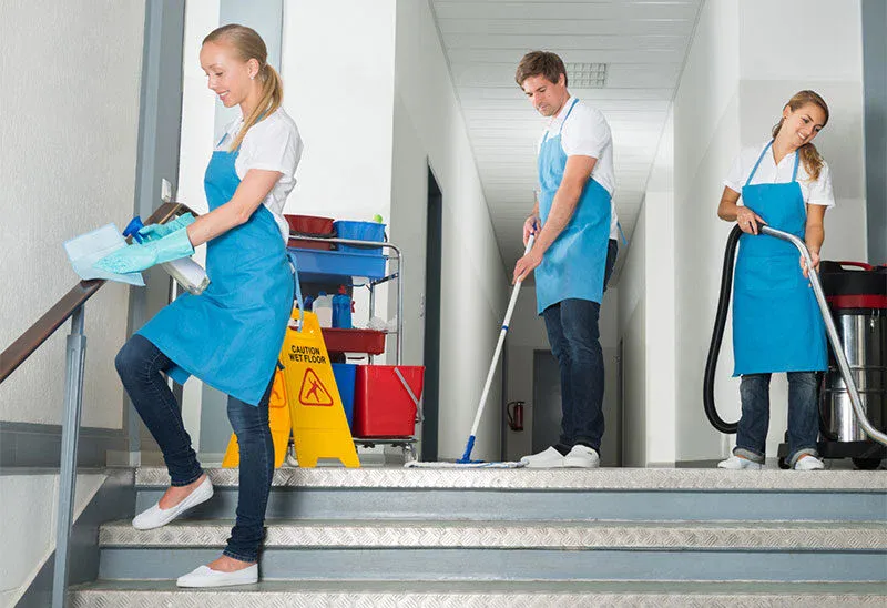 Three people in blue aprons and white shirts are cleaning a hallway and stairs. One wipes a stair railing, another mops the floor, and the third vacuums. Cleaning equipment and a caution wet floor sign are visible.