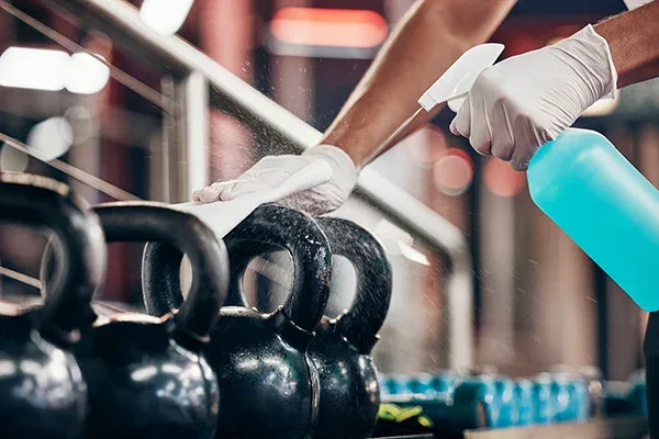 A person wearing white gloves sprays disinfectant and wipes down black kettlebells in a gym, focusing on cleanliness and sanitation.