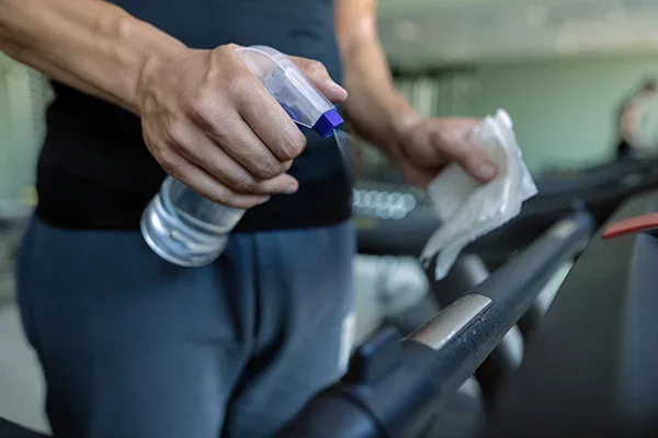 A person cleaning gym equipment with a spray bottle and a cloth, focusing on wiping down a treadmill handle.