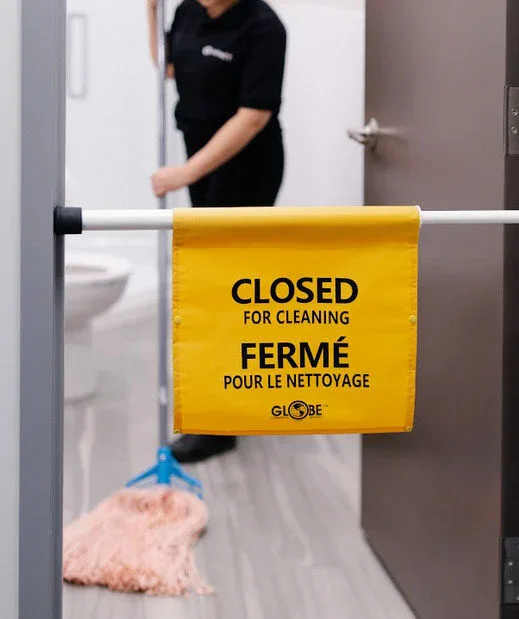 A yellow Closed for Cleaning sign hangs on a bar across a restroom door, with a person mopping the floor inside. The sign, in English and French, reflects the high standards of impact commercial cleaning services in Canada.