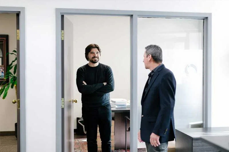 A man in a black sweater stands in an office doorway, arms crossed, smiling at another man in a suit who is standing in the hallway. Their discussion highlights the positive effect of impact commercial cleaning services on workplace environments.