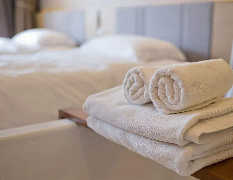 Two neatly rolled white towels rest atop folded towels on a wooden surface in a clean, modern hotel room, reflecting the standards of impact commercial cleaning services with a blurred bed and pillows in the background.