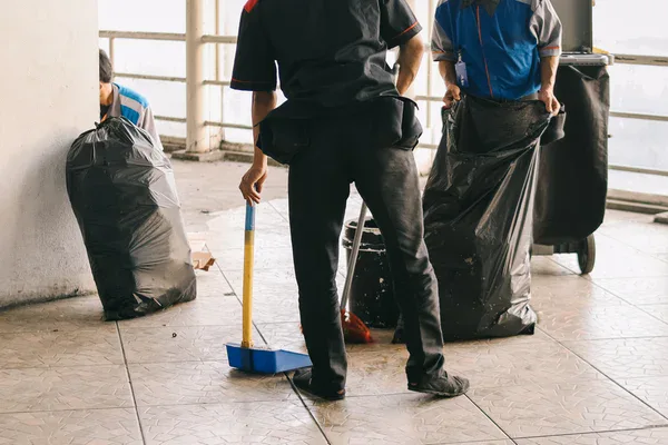 Three people are cleaning a tiled floor; one holds a dustpan and broom while the others handle large black trash bags. A railing is visible in the background.