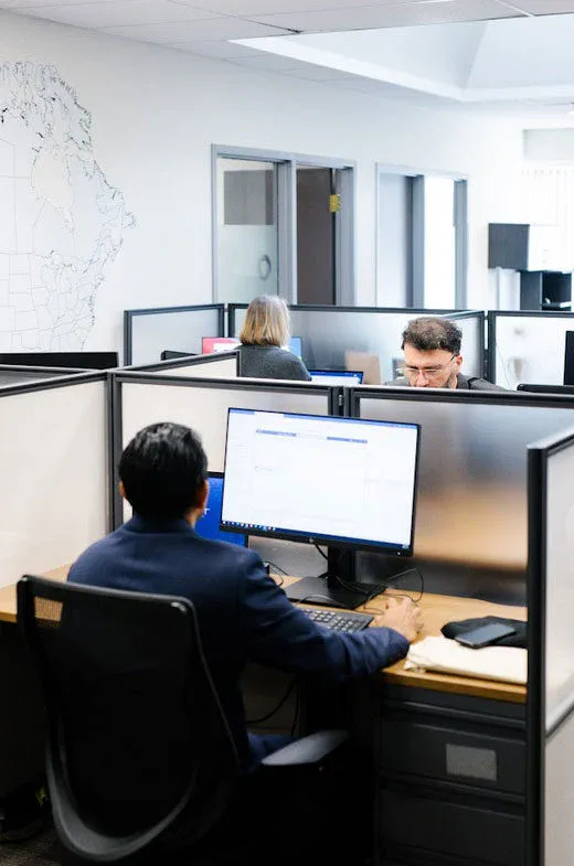 Three people work at individual cubicles in a modern office, maintained by commercial cleaning Canada. In the foreground, a man uses a computer, while two others are visible in the background—one facing forward and one with their back to the camera.