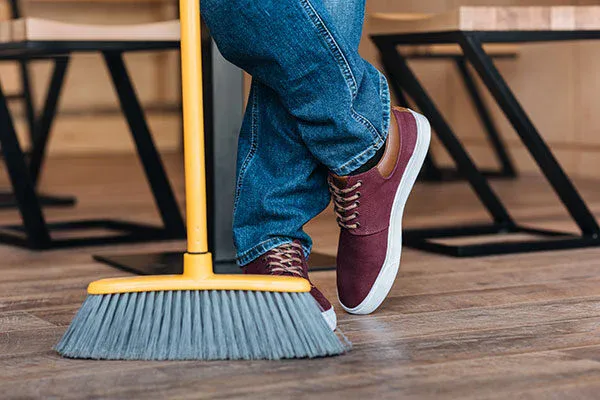 Person wearing blue jeans and maroon sneakers stands with crossed legs, holding a yellow broom on a wooden floor, with chairs and tables in the background—showcasing the impact of commercial cleaning services in Canada.