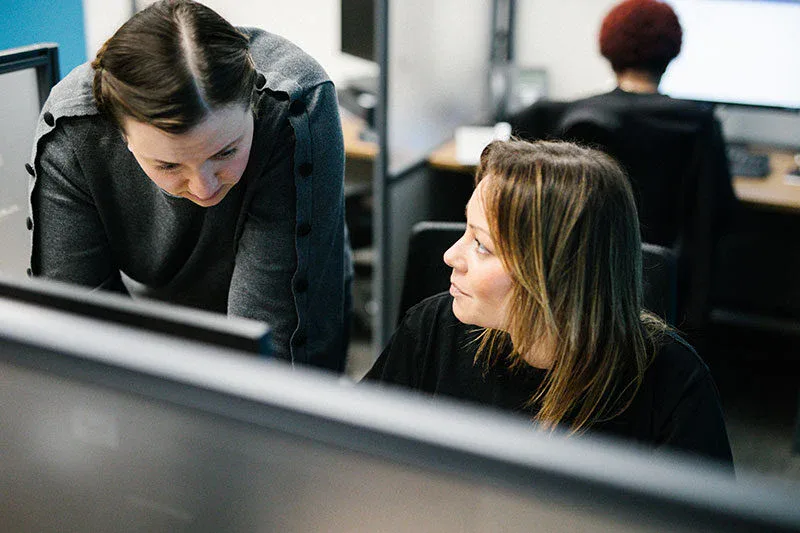 Two women in an office setting have a focused conversation at a desk, highlighting the impact commercial cleaning services offer in maintaining professional environments, with computers and another person working in the background.