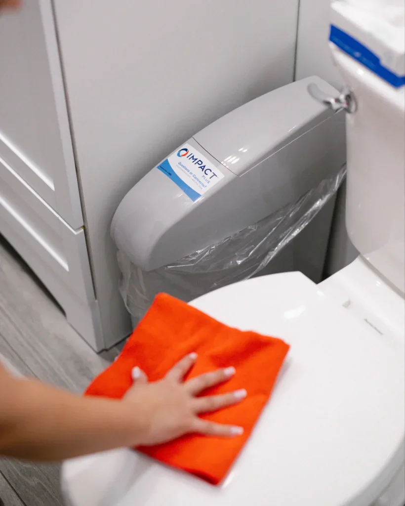 A person cleans a white toilet seat with an orange cloth. The toilet tank has a plastic-lined cover labeled IMPACT, reflecting impact commercial cleaning services. The bathroom floor is light wood, and white cabinets are visible in the background.