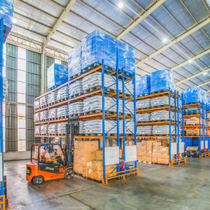 A worker drives an orange forklift past tall racks stacked with pallets, boxes, and goods in a spacious, brightly lit warehouse with a high ceiling.