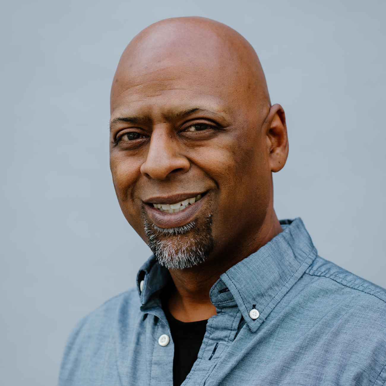 A bald man with a goatee and a slight smile is wearing a blue button-up shirt over a black t-shirt. He is standing against a plain, light gray background.