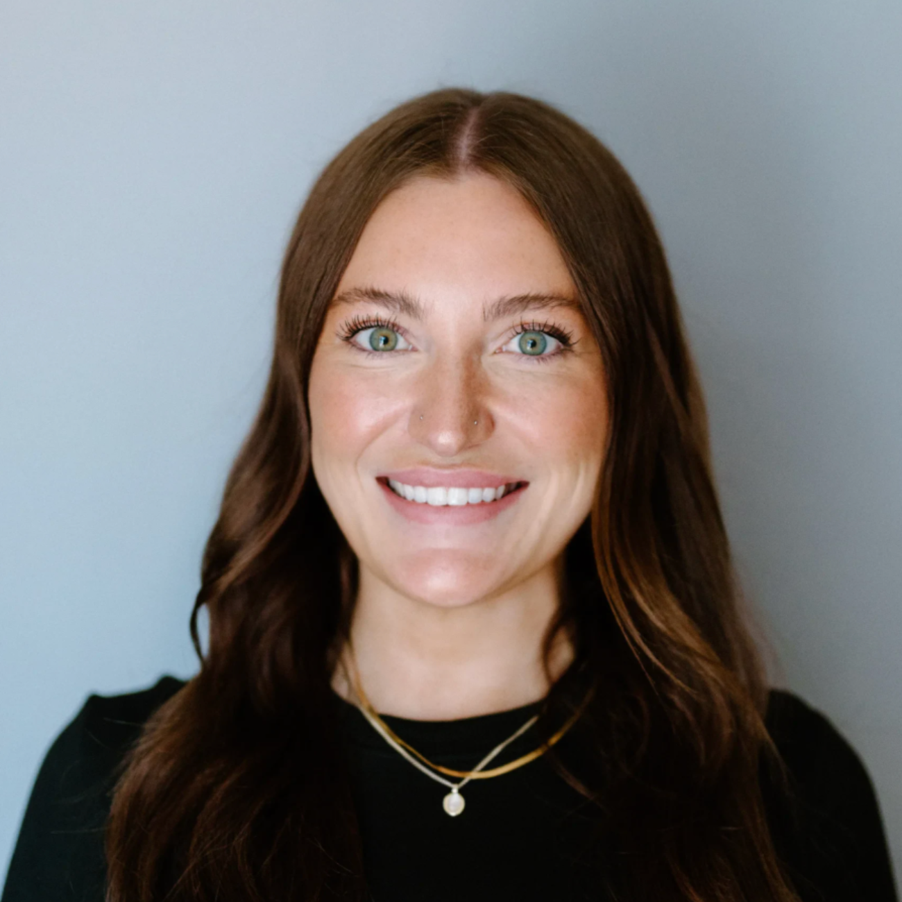 A woman with long brown hair and green eyes smiles at the camera. She is wearing a black top and two gold necklaces, standing in front of a plain light gray background.