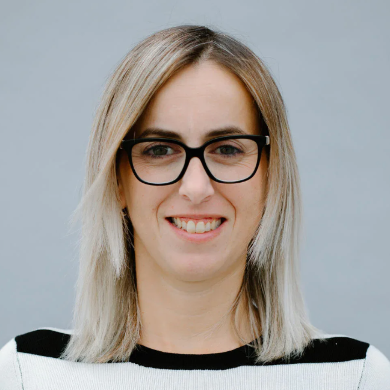 A person with straight, shoulder-length blonde hair and black glasses smiles at the camera, wearing a white and black top, with a plain gray background.